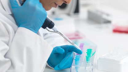 A woman in a lab coat wearing blue gloves is holding a pipette and filling a sample vial symbolising pharmaceutical impurity analysis services in a laboratory setting.