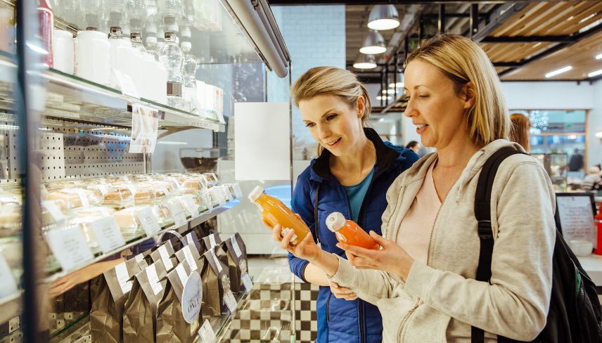 Two women comparing bottled drinks while shopping at a grocery store shelf.